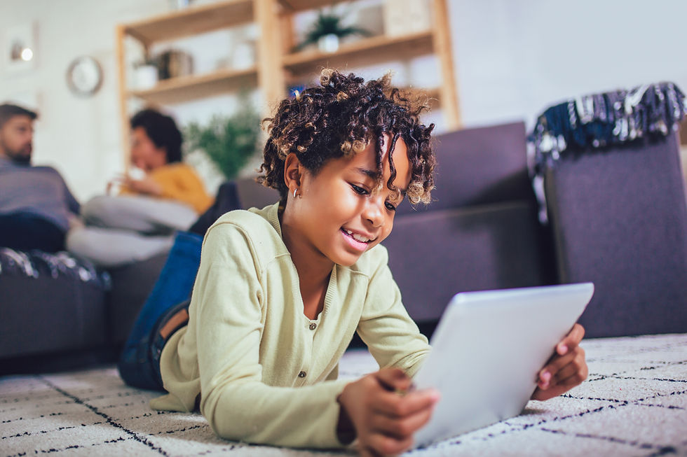A child is lying on the floor holding a tablet while the parents are in the background sitting on the couch talking.