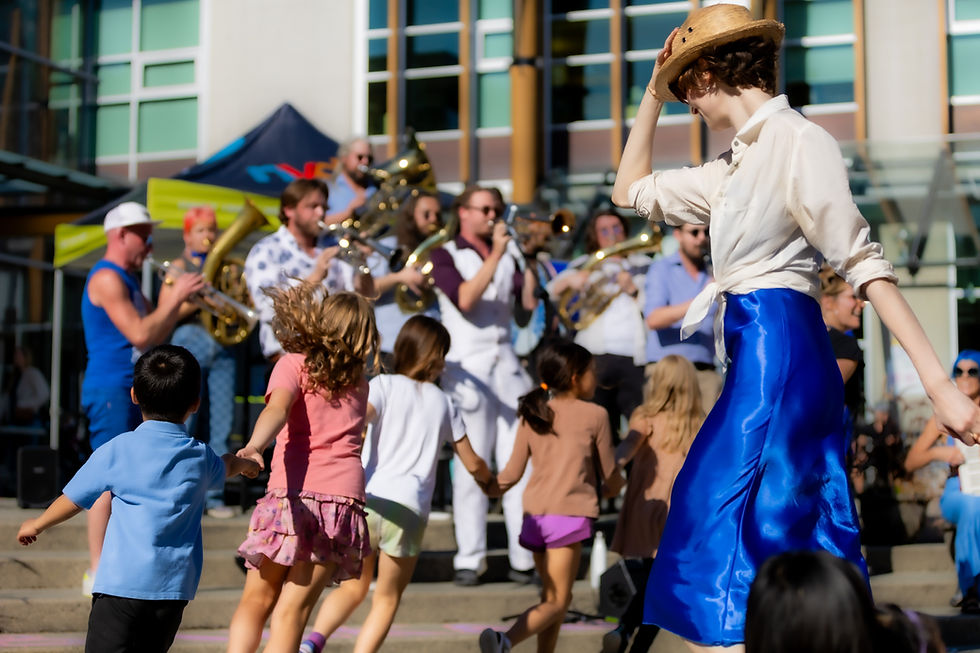 An outdoor scene with a group of children and adults participating in a lively event. In the foreground, a woman wearing a straw hat and blue skirt leads dancing with children. A brass band is in the background playing music.
