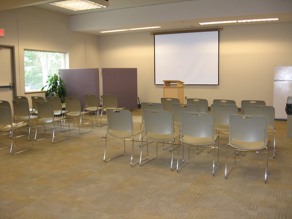 A beige meeting room with four rows of chairs facing a lecturn in front of a projector screen. There is a small window on the wall to the left.