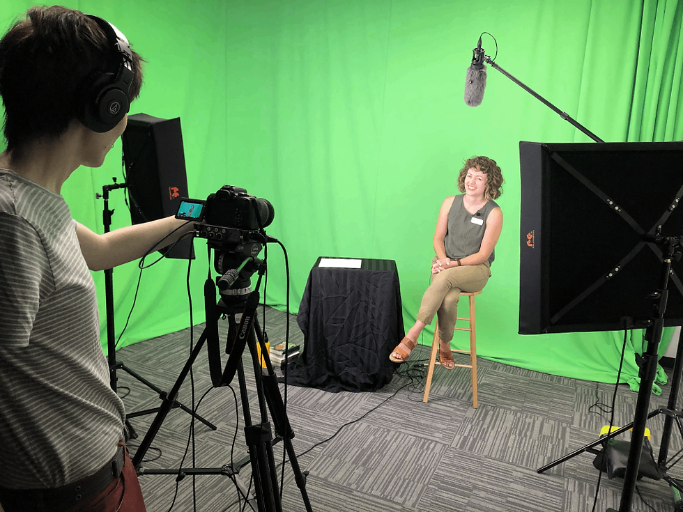 A person sitting on a stool crossed legged acing a camera with a camera person. The room is covered in green fabric and there is lightbox and boom mic visible.
