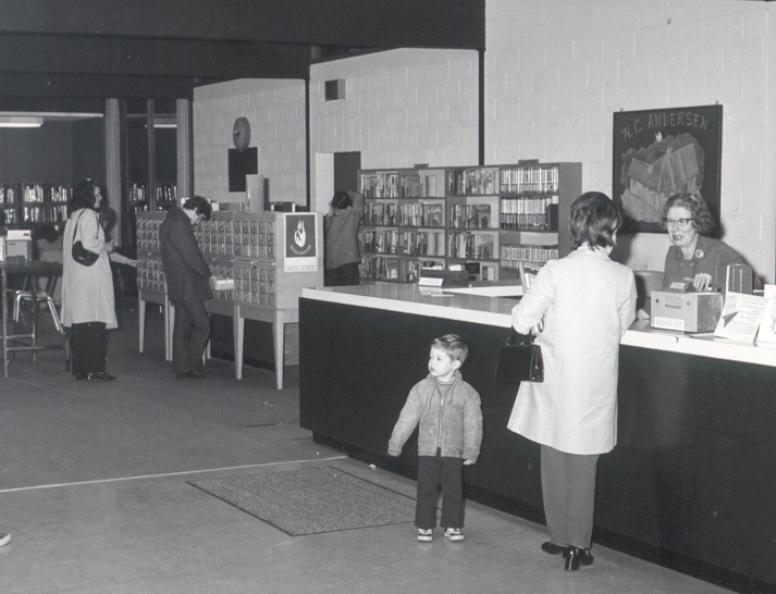 A black and white picture of the Capilano Branch in 1969. A patron with their child stands speaking with library staff at the circulation desk. Patrons in the background browse card catalogue drawers.