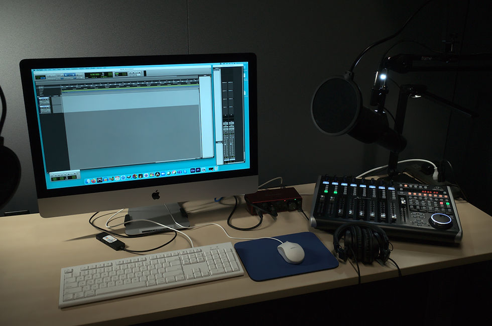 Tabletop filled with audio equipment including a Mac computer, headphones, microphone, and a audio mixer. The computer is turned on is showing audio editing software.
