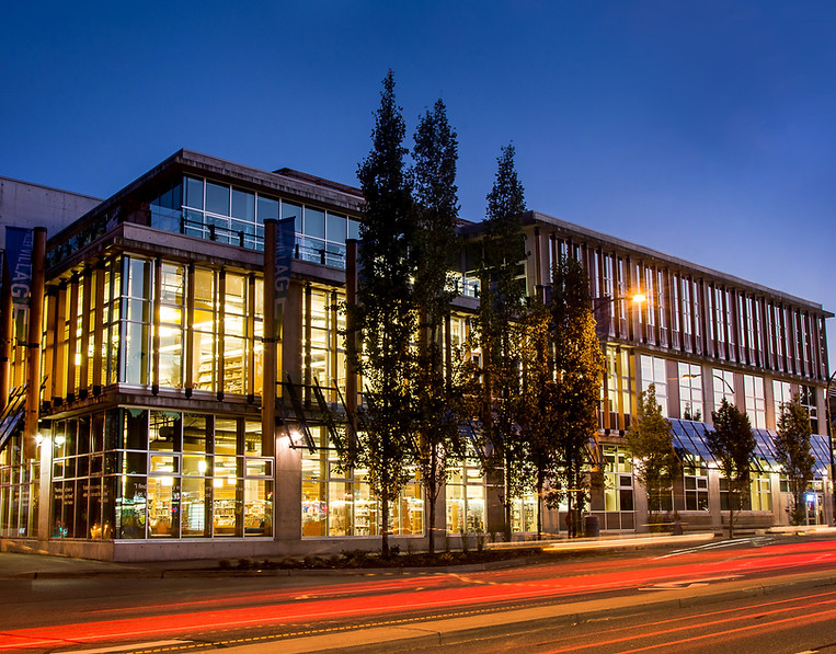 A picture of the corner of Lynn Valley Library at dusk. Lights are on in the library and a dark blue sky can be seen behind the library.