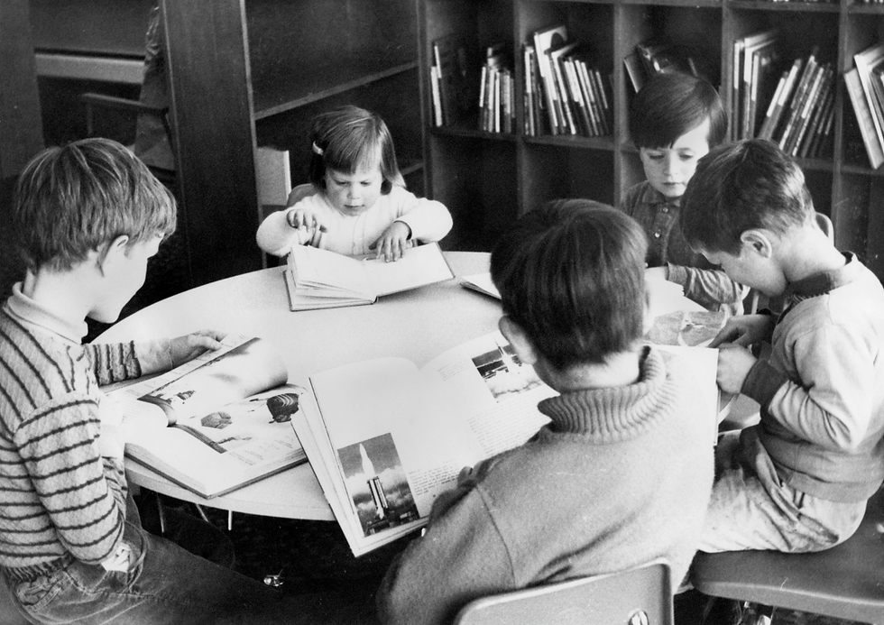 A black and white photo of children sitting at a table reading books at the old Deep Cove Library, circa 1970.