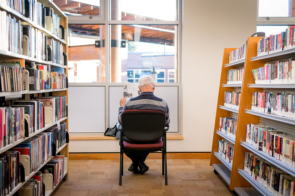 A man sits on a chair in the library, between two book shelves. He reads a newspaper facing the library window.